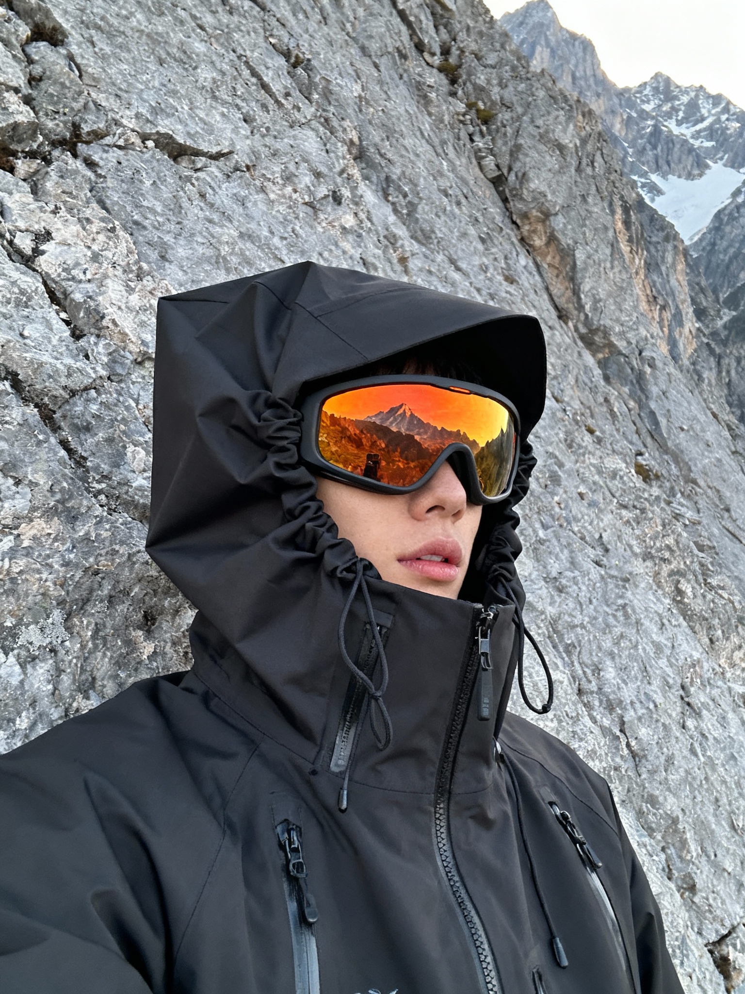 A naturally posed side profile of an individual standing relaxed in front of a textured rugged rock wall within a mountainous terrain. The person wears a matte black technical waterproof hardshell jacket with an oversized hood fully raised, featuring angular shaping and cinch cords creating a sculptural, structured form that closely envelops the head. The jacket shows taped seams and waterproof zippers, highlighting its high-performance, utilitarian design. The individual sports high-contrast, orange-tinted wraparound goggles that vividly reflect the surrounding natural alpine landscape, partially obscuring the face so only the nose and part of the lips are visible beneath the hood and eyewear. Soft, diffused, indirect daylight gently illuminates the scene, emphasizing the rich textures of the waterproof fabric and the coarse rocky surfaces behind. The casual, spontaneous framing includes a subtle, slightly tilted angle, reinforcing the authentic candid iPhone photography aesthetic. This hyper-realistic, cinematic image conveys a calm, tactical, futuristic outdoor vibe within a rugged mountain setting, seamlessly blending minimalist mountain utility with cyberhiker fashion.