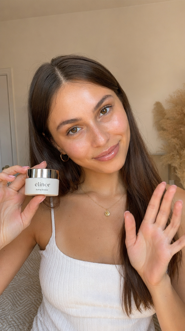 Authentic UGC selfie photo of a young woman in her early twenties, light olive skin with a fresh dewy glow, long straight brown hair parted in the middle falling past her shoulders, warm brown eyes, soft natural brows, glossy nude lips, subtle highlight on cheekbones. Wearing a white ribbed spaghetti strap top, delicate gold pendant necklace at the collarbone, small gold hoop earrings, light pink manicure. She is holding a small skincare jar up to her left side at chest level — the jar has a white matte body with a polished chrome silver twist lid, minimalist serif logo reading "elinor" on the front, smaller text underneath reading "Sunray bronzer". Her right hand is raised palm out toward camera in a casual wave gesture, slight head tilt, soft confident smile, looking directly into the lens. Soft neutral bedroom interior background, slightly out of focus warm beige wall. Soft natural window light from front and slightly above, warm daylight tone, gentle falloff. Dewy luminous skin with real visible pores, no heavy retouching. Shot on iPhone front camera, medium close up, head and chest in frame, eye level very subtle low angle, shallow depth of field with soft background blur. Warm neutral color palette, cream, beige, peach skin tones, hint of golden warmth, clean lifestyle aesthetic. Vertical portrait orientation. No play button, no video player UI, no timestamp, no watermark, no text overlays.