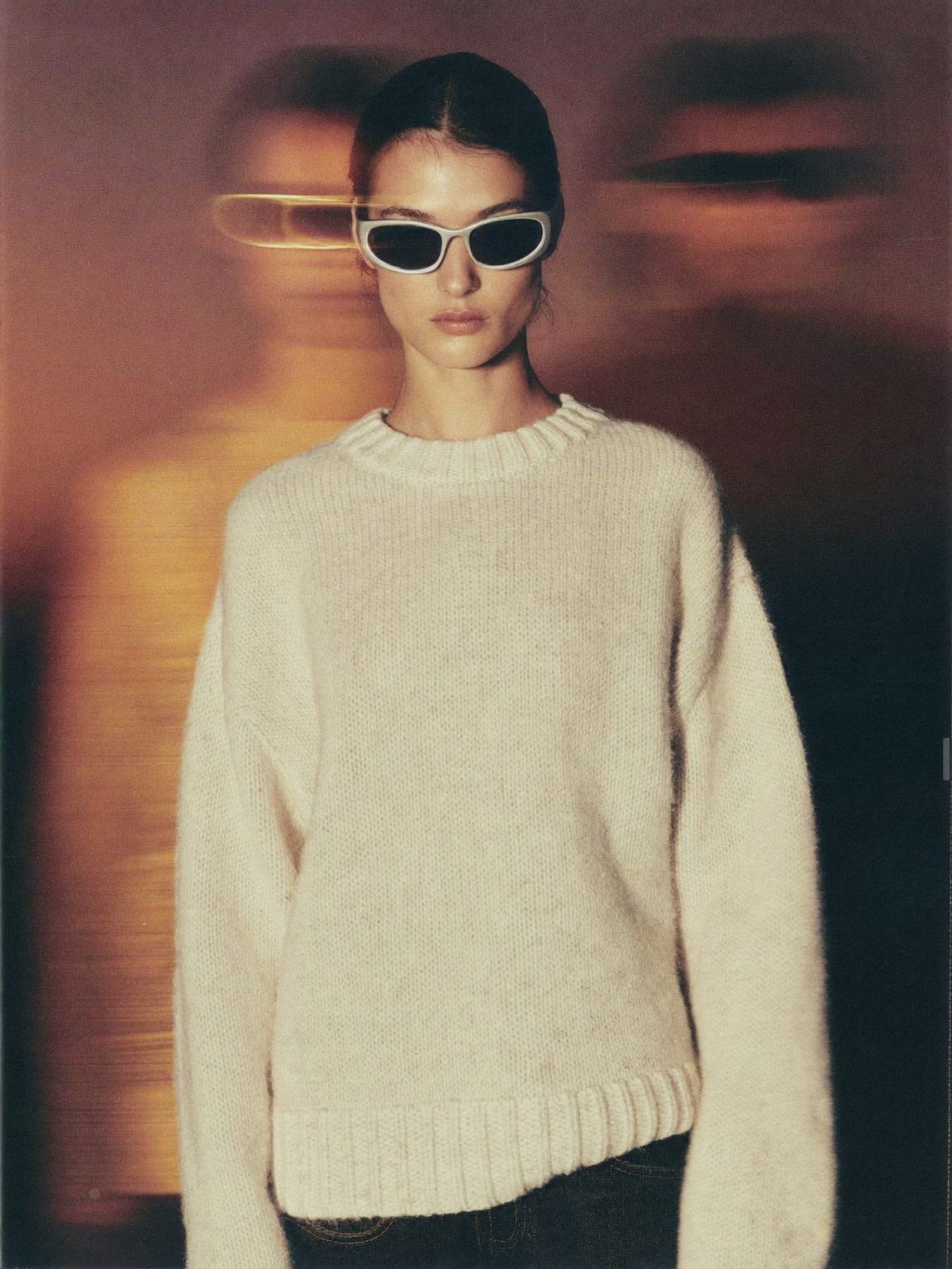 Editorial fashion photograph, long exposure with intentional camera motion blur. A young woman in a cream oversized knit and silver wraparound sunglasses stands still and sharp in the center while ghosted, smeared double exposures of two other people drift behind her. Warm dusk light, amber and rose sunset glow smeared horizontally, deep mauve sky, soft film grain, shallow depth of field. Muted, desaturated palette of cream, amber, rose, charcoal. No green tones, no text, no logos.